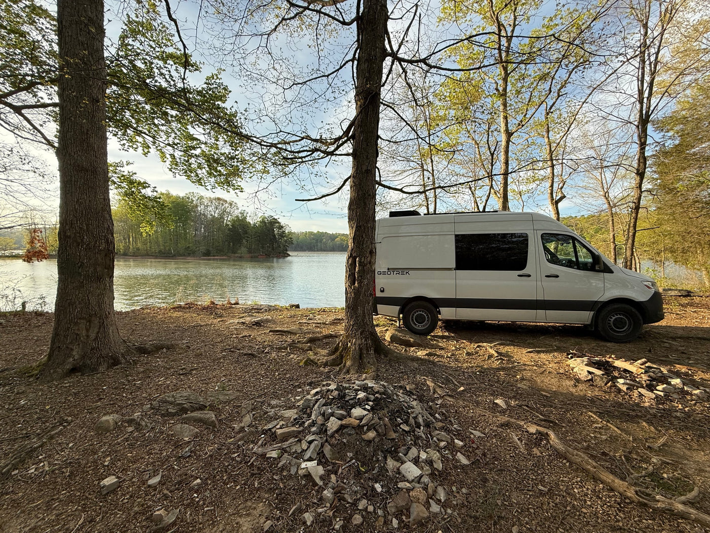 Lakeside RV rental: A white camper van parked on a dirt campsite surrounded by trees, offering peaceful nature views. — photo 8
