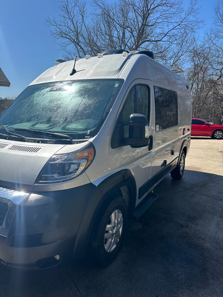 Silver camper van, ready for your RV rental journey, shines in the sun. Parked outdoors with bare trees and a red car in distance.