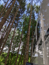 Tall trees tower over a gleaming rv rental, its side reflecting the lush green canopy and blue sky in a peaceful campsite.