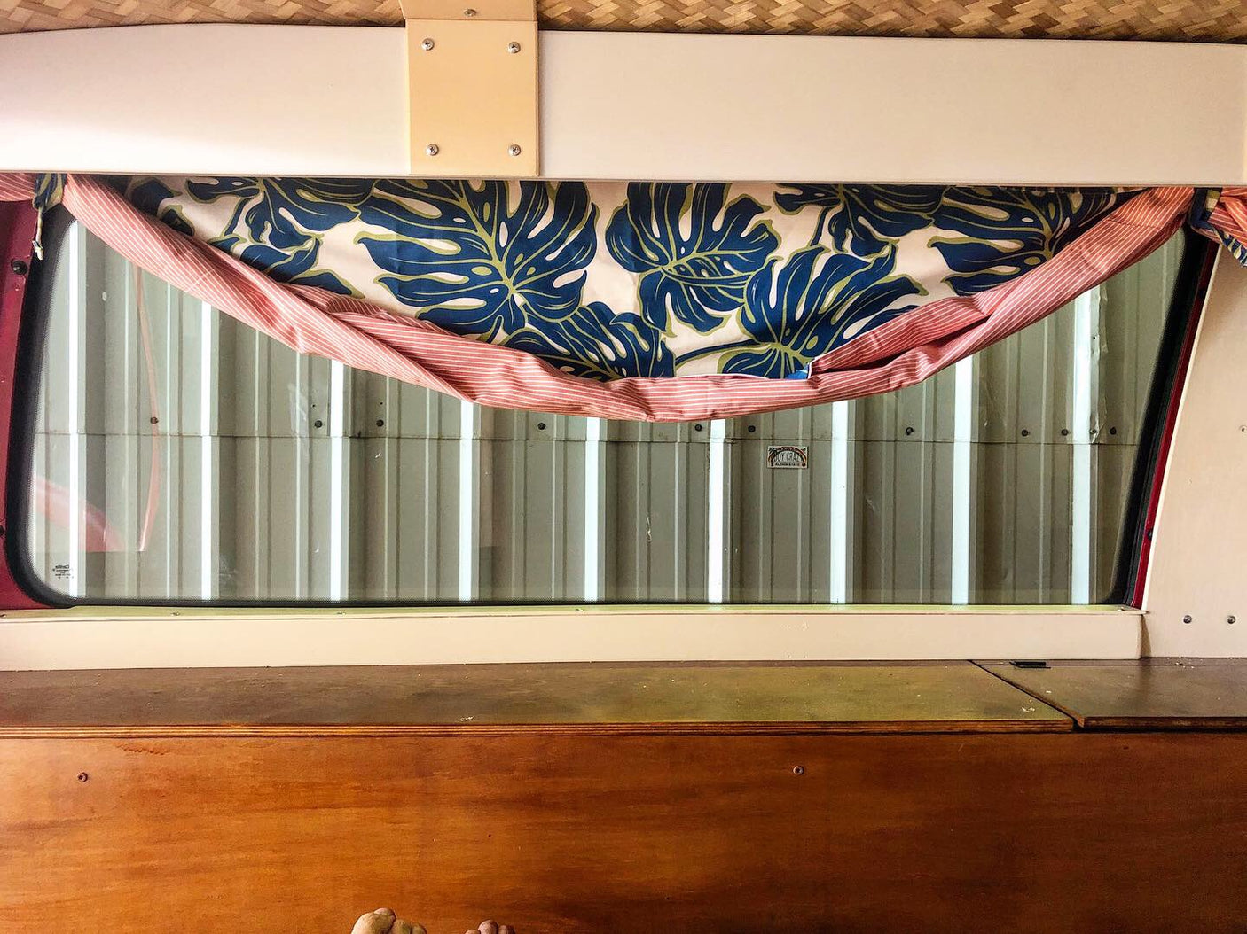 Cozy RV rental interior with a tropical leaf curtain, offering a window view of a corrugated wall and a wooden bed.
