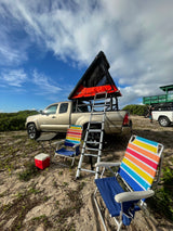 Scenic beach camp setup! Tan truck with a rooftop tent, ladder, and colorful chairs. Consider an rv rental for adventure.