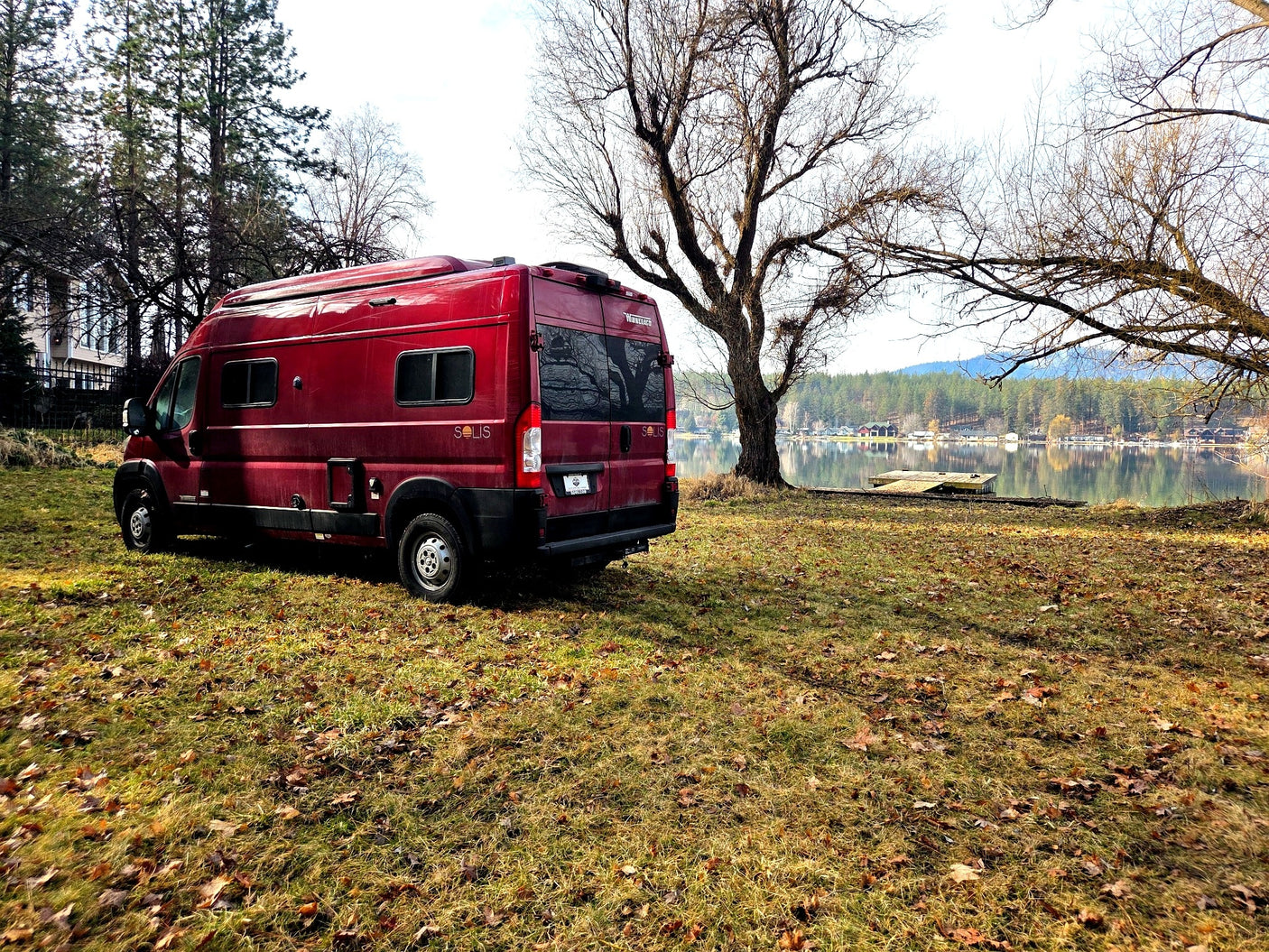 Red Winnebago Solis RV rental parked on grassy ground by a serene lake, with bare trees and forested hills in the distance.