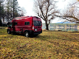 Red Winnebago Solis RV rental parked on grassy ground by a serene lake, with bare trees and forested hills in the distance.
