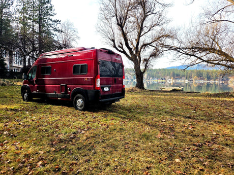 Red Winnebago Solis RV rental parked on grassy ground by a serene lake, with bare trees and forested hills in the distance.