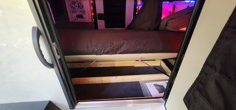 Inside an RV rental: bed lifted with gas struts to reveal spacious under-bed storage. "Don't Wake the Bear" sign on wall.