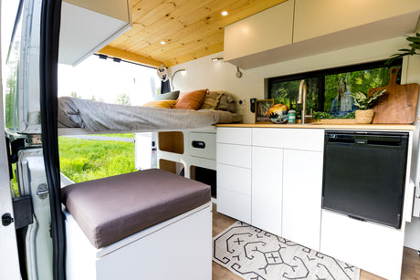 Cozy RV rental interior: raised bed, sleek kitchen with sink & fridge, white cabinets, and scenic window views.