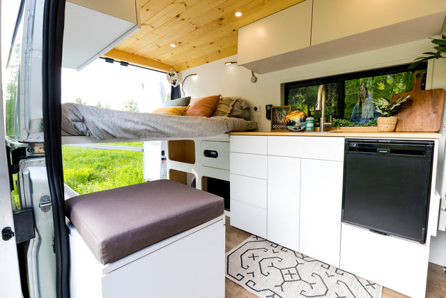 Cozy RV rental interior: raised bed, sleek kitchen with sink & fridge, white cabinets, and scenic window views.