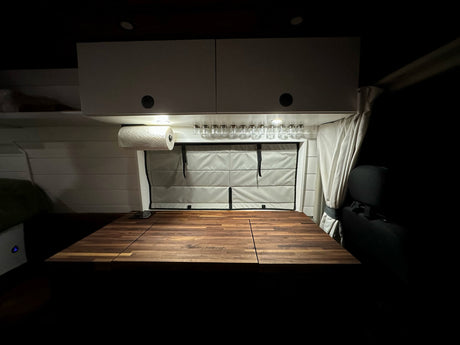 RV rental's compact kitchen area: lit cabinets, paper towels, spice jars, and a folding wooden table setup.