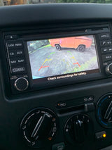Car's backup camera shows a vintage orange RV rental. Dashboard controls aid safe reversing in a detailed car interior.