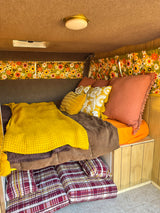 Cozy, retro RV rental interior featuring vibrant floral curtains, yellow knit blanket, plush pillows, and wood paneling.