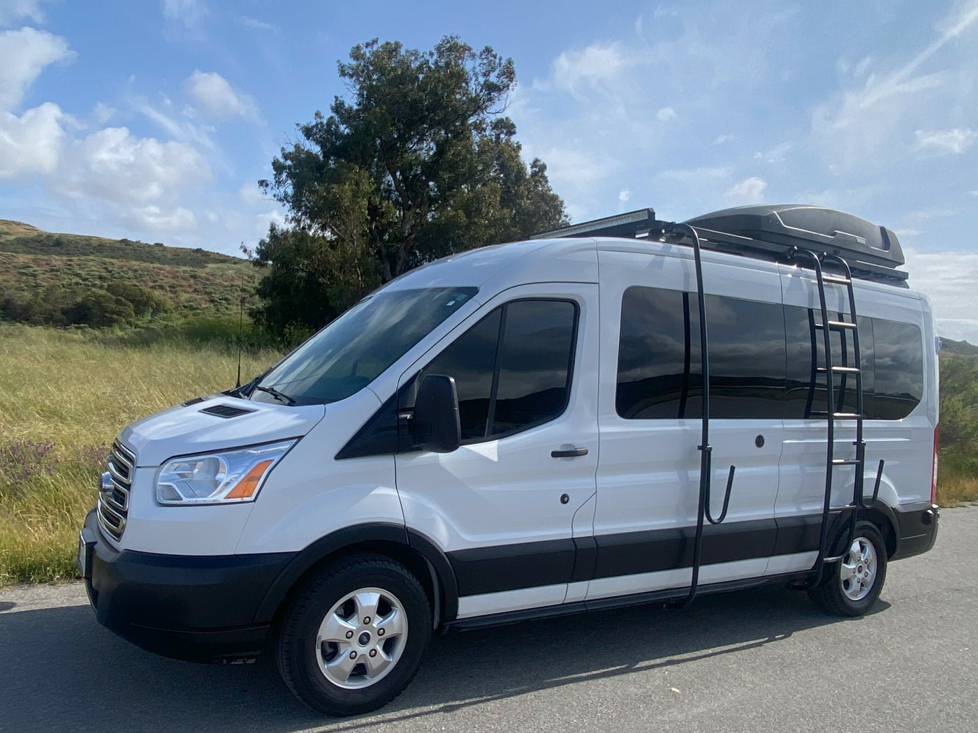 White Ford Transit RV rental van, customized with a roof rack, cargo box, and ladder, parked roadside against a green hill. — photo 4