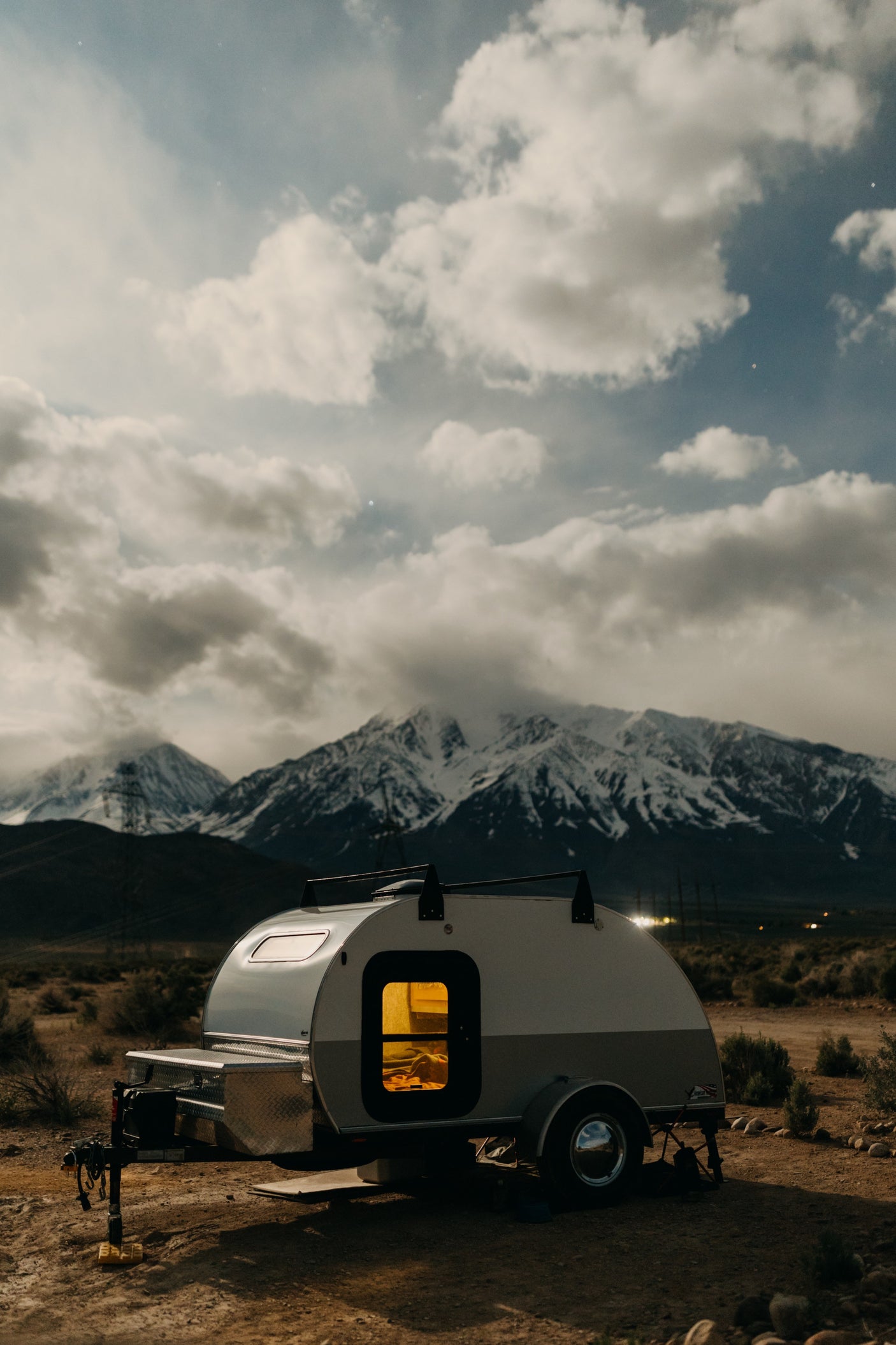 A cozy teardrop RV rental glows at dusk, parked in a desert landscape with majestic snow-capped mountains under a cloudy sky.