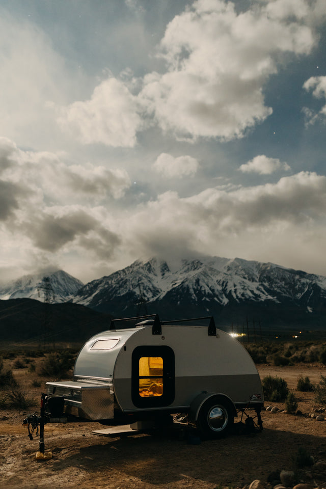 A cozy teardrop RV rental glows at dusk, parked in a desert landscape with majestic snow-capped mountains under a cloudy sky.