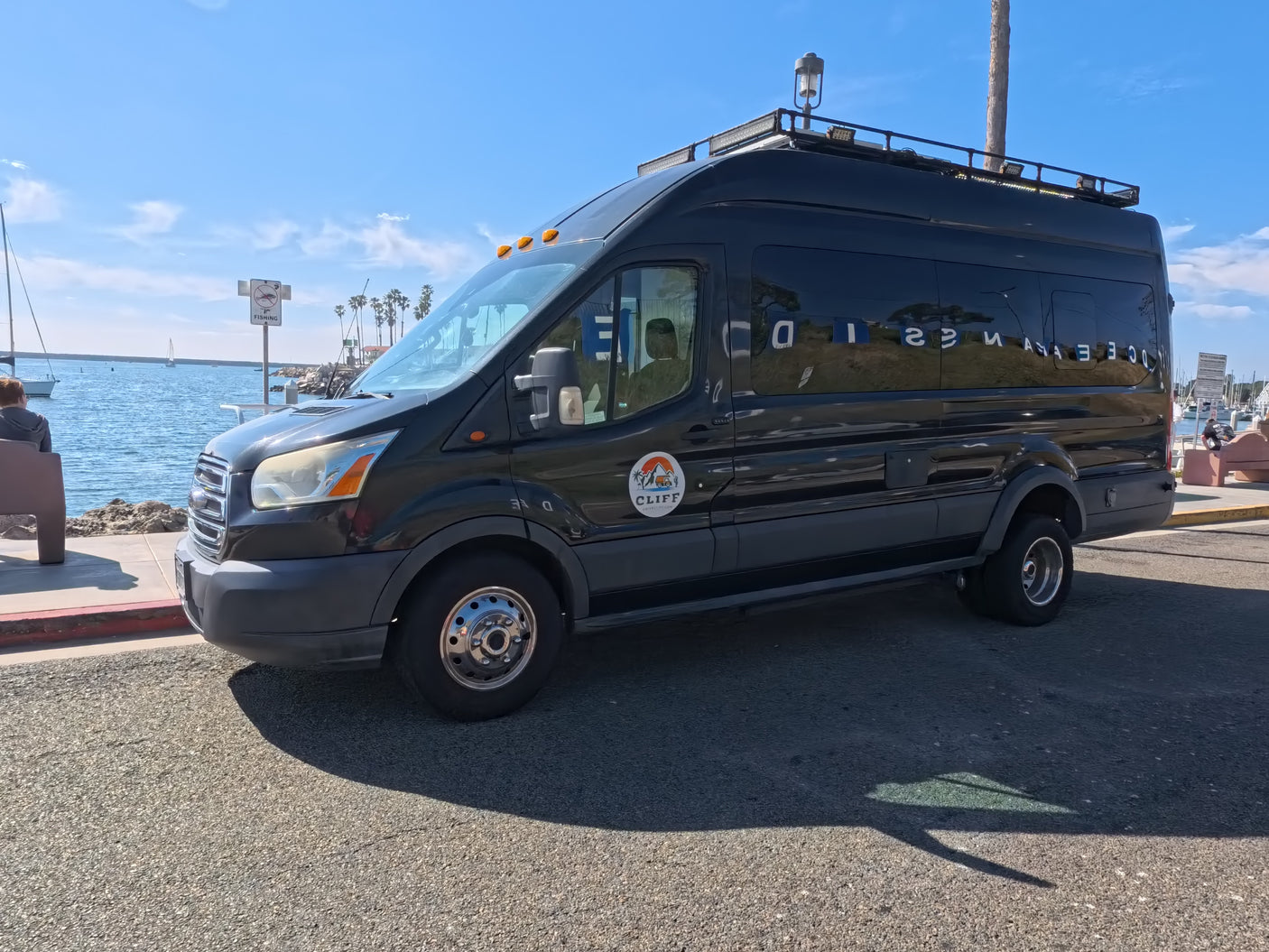 A black rv rental van (CLIFF logo) is parked by an ocean marina with boats & palm trees under a sunny sky.
