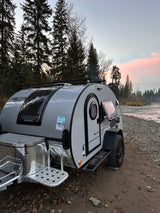 Teardrop RV rental parked by a peaceful river at sunset, surrounded by tall trees. Ideal for a nature camping trip.
