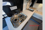 RV rental interior: galley with a 2-burner stove, sink, and counter. Light-colored passenger seating visible.