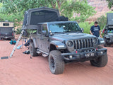 RV rental ready: Grey Jeep Gladiator with rooftop tent, a truck camper, and another rig set up for overlanding on red dirt.