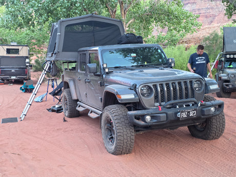 RV rental ready: Grey Jeep Gladiator with rooftop tent, a truck camper, and another rig set up for overlanding on red dirt.