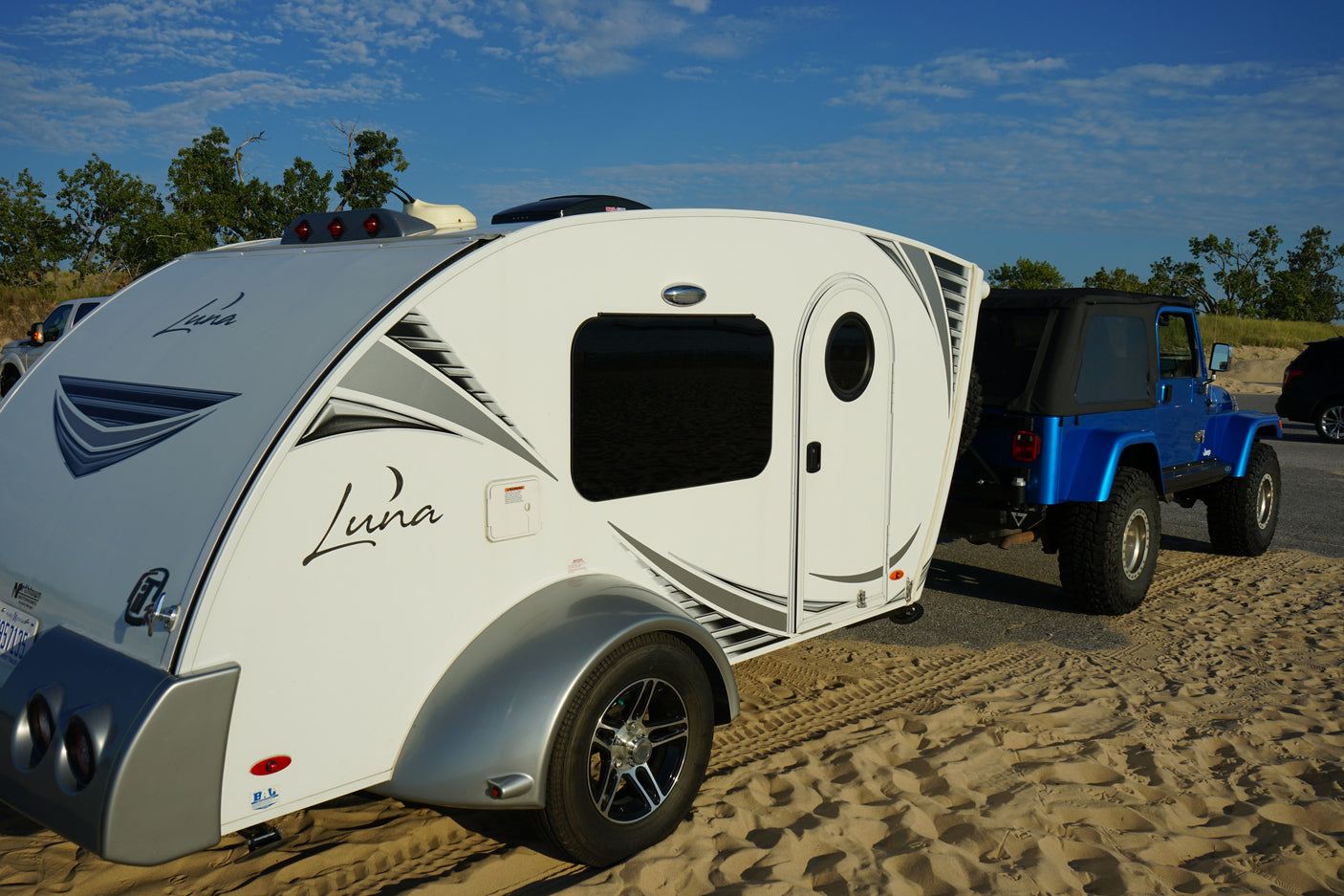 A white Luna teardrop RV rental hitched to a blue Jeep, ready for beach or sand dune camping.