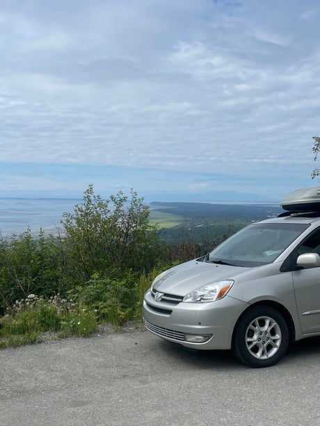  parked at a scenic overlook with a bay view.|View from rear of a minivan converted for car camping or an RV rental. A comfortable bed with white bedding and a colorful crochet blanket fills the back.|Silver minivan with roof box at a scenic overlook