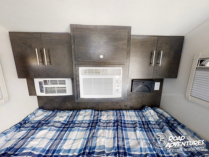 Cozy RV rental bedroom with a plaid bed, overhead cabinets, and dual AC units, perfect for comfortable road adventures.