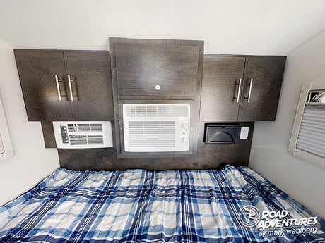 Cozy RV rental bedroom with a plaid bed, overhead cabinets, and dual AC units, perfect for comfortable road adventures.