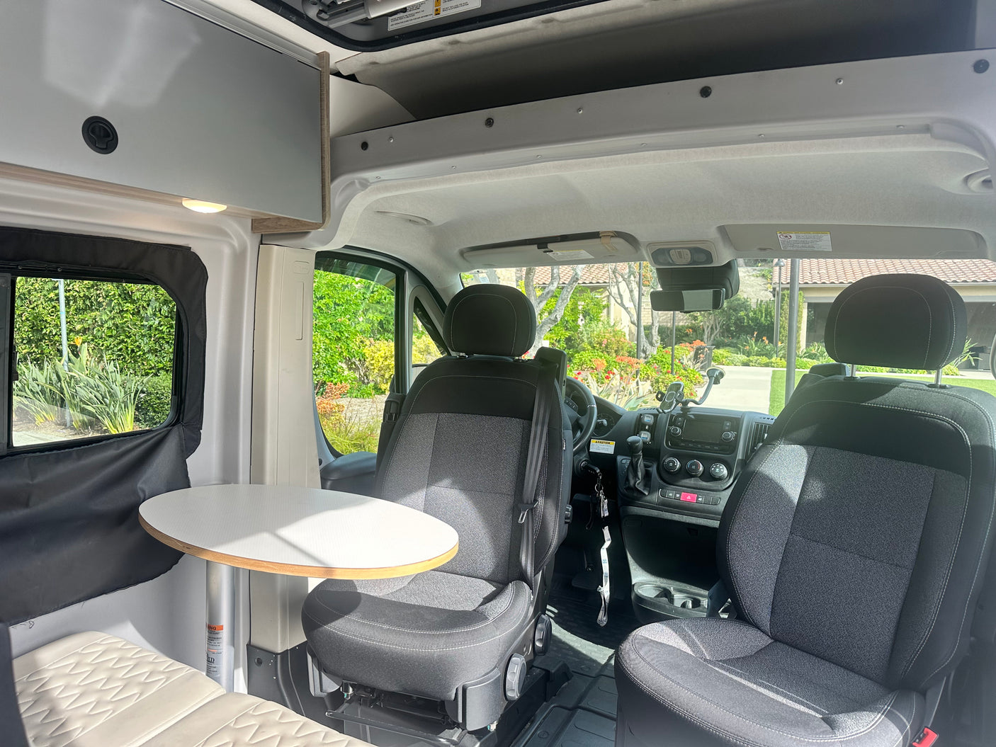 View of an RV rental camper van's interior: front seats, dashboard, dining table, & overhead storage.