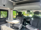 View of an RV rental camper van's interior: front seats, dashboard, dining table, & overhead storage.