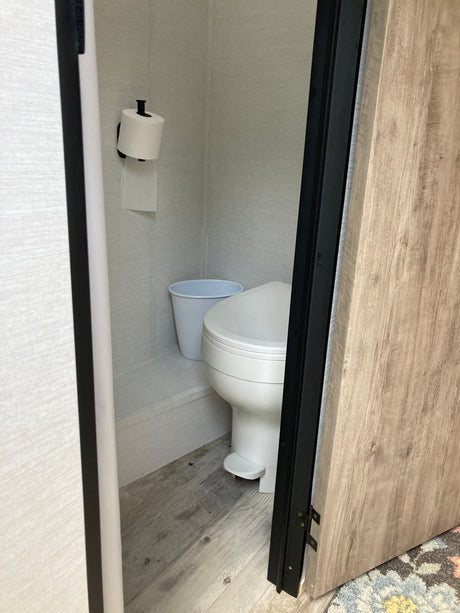 RV rental: A compact bathroom featuring a toilet, toilet paper roll, and white bucket on light wood flooring.