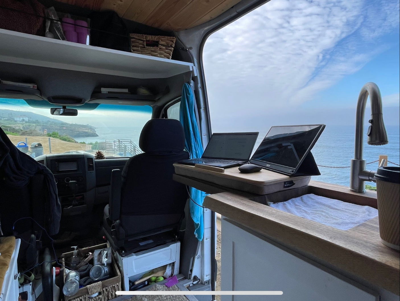 Inside an RV rental, a mobile office with laptop & monitor overlooks the ocean. A sink & faucet are to the right.