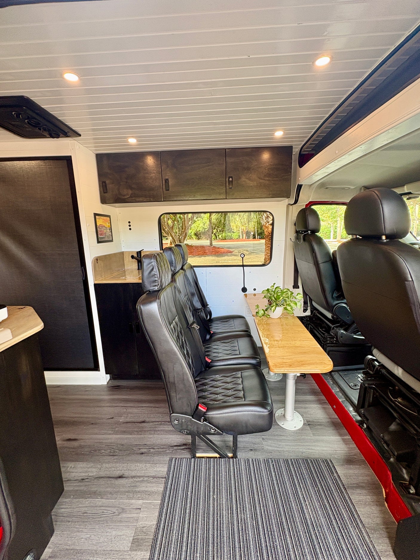 Inside an RV rental camper van: black leather seating, a wooden table with a plant, and a kitchenette, with a view of trees.