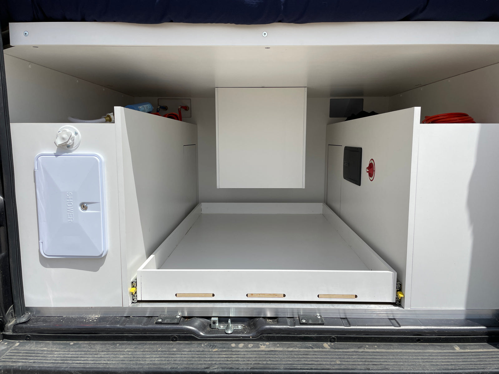 Inside an RV rental: rear utility area with pull-out storage, outdoor shower, and visible plumbing/electrical connections.