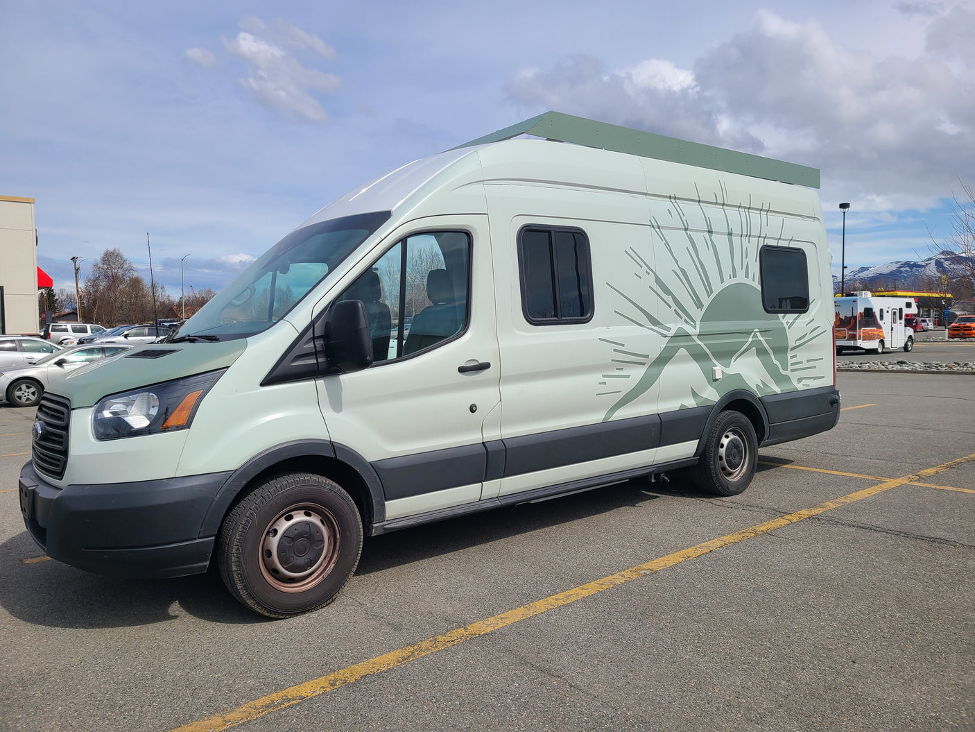 RV rental: Light green camper van with mountain graphic and elevated roof, parked in a lot with snowy mountains.