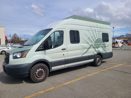 RV rental: Light green camper van with mountain graphic and elevated roof, parked in a lot with snowy mountains.