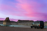 Black RV rental van by a serene lake at sunset, showcasing vibrant purple and pink skies above rugged rock formations.