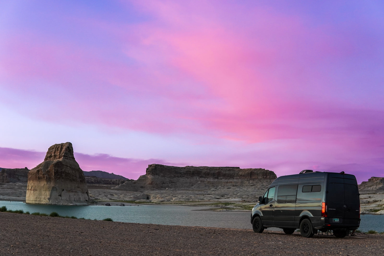 Black RV rental van by a serene lake at sunset, showcasing vibrant purple and pink skies above rugged rock formations.