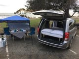 Minivan camper setup for beach camping, a great alternative to rv rental. Features a bed, canopy, & outdoor kitchen.