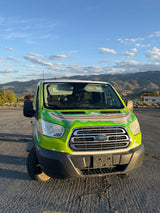 Front view of a bright green Ford Transit RV rental van, parked outdoors with mountains and a blue, cloudy sky.