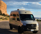 RV rental: Beige Mercedes Sprinter camper van on a desert road, by a large rock formation.