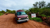 Maroon rv rental van on a dirt road in Hawaii. A sign prohibits driving on the beach, overnight parking, and camping.