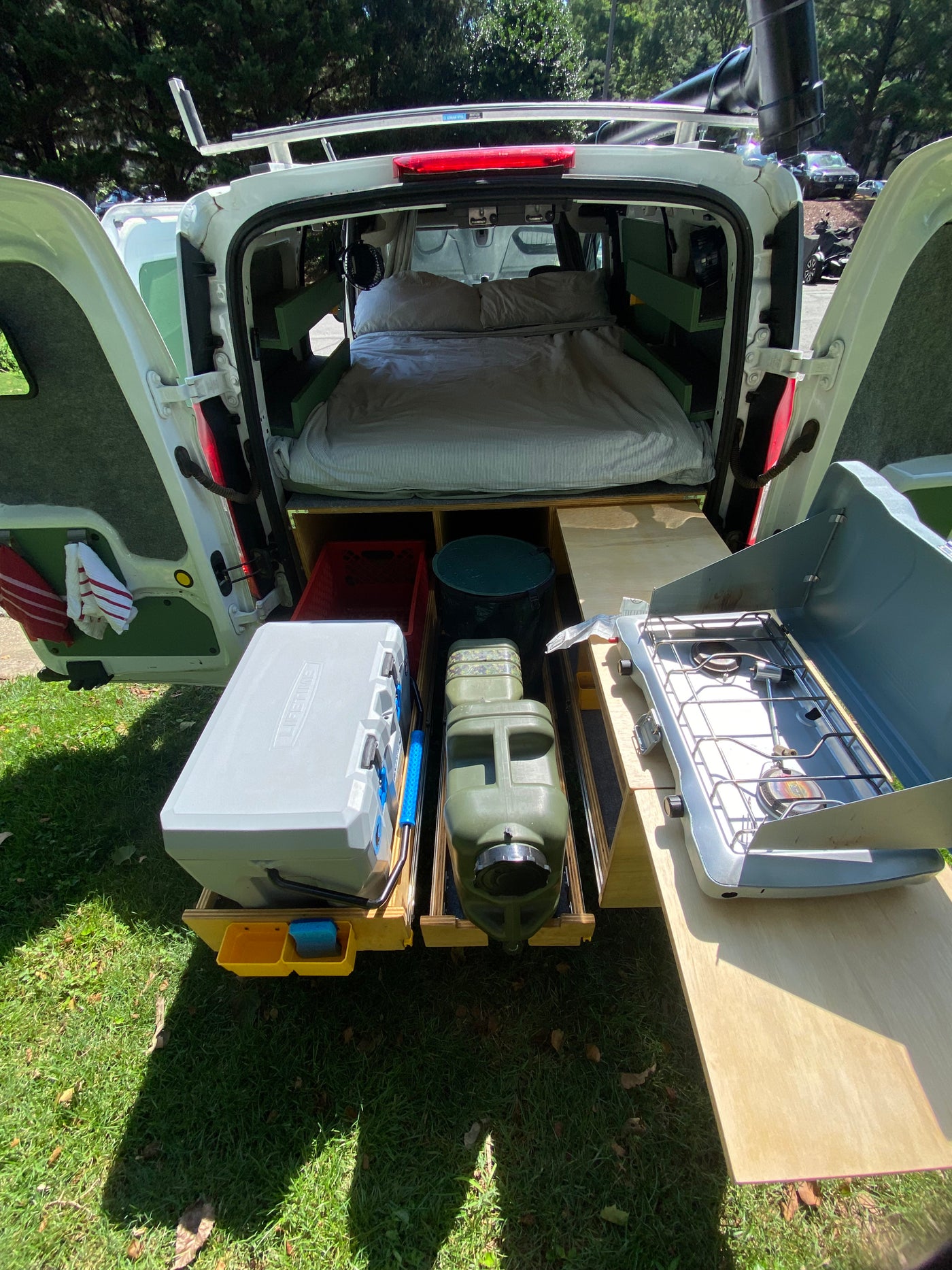 Rear view of a fully equipped camper van conversion for **rv rental**. Features a bed, pull-out cooler, water jugs, & camp stove for outdoor living. — photo 7