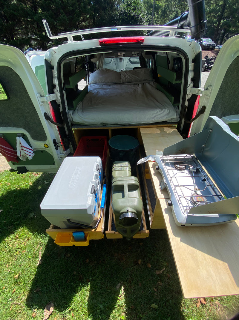 Rear view of a fully equipped camper van conversion for **rv rental**. Features a bed, pull-out cooler, water jugs, & camp stove for outdoor living. — photo 7