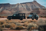 Black Jeep Gladiator and an overland trailer in a rugged desert landscape with red mountains. Ideal RV rental for exploring.