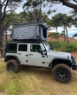 Silver Jeep Wrangler with rooftop tent, parked amidst pines & red hills. An RV rental option for overlanding.