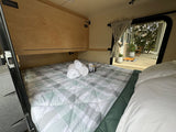 Cozy RV rental interior: A compact bed with plaid bedding, fresh towels, and an open door leading to an inviting patio.