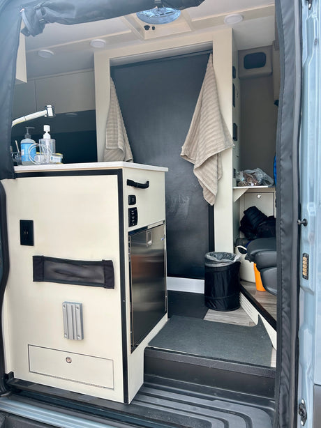 View inside an rv rental's compact camper van, with kitchenette, sink, fridge, storage, and open entry.