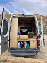 This converted van's open rear reveals a cozy interior & smart storage, perfect for an RV rental adventure on sandy terrain.