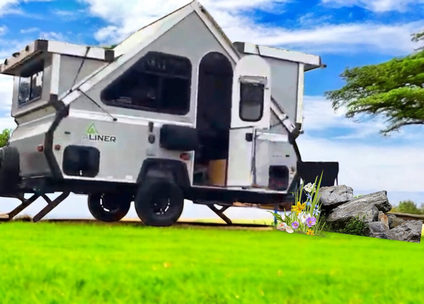 An A-Liner A-frame RV rental is set up on a vibrant green field under a blue sky, with rocks and flowers nearby.
