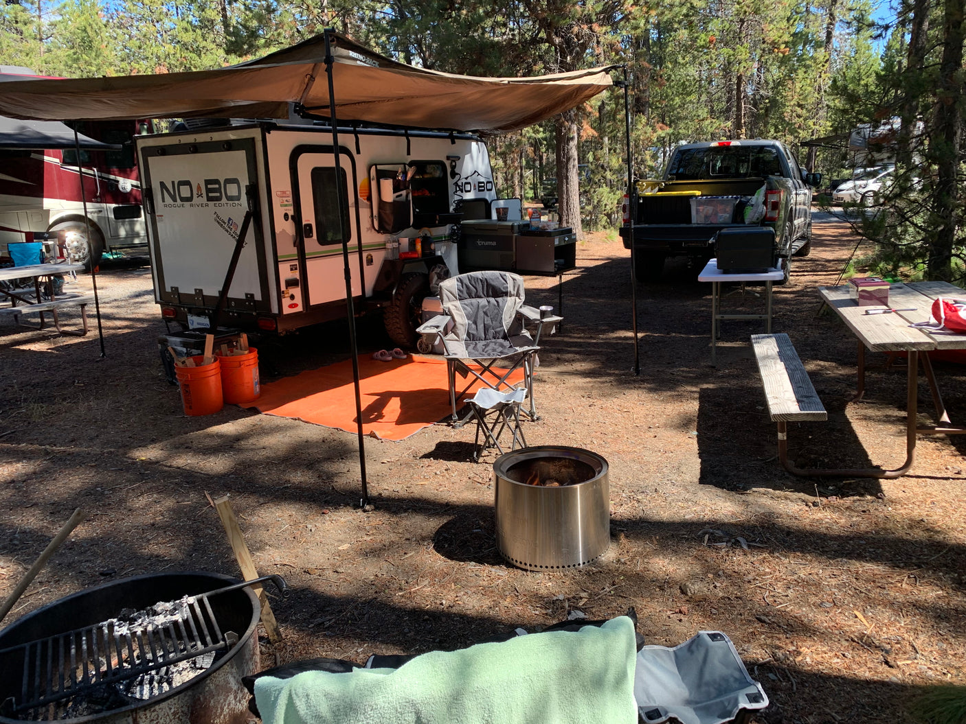 Relaxing RV rental campsite with a NO BO travel trailer, truck, fire pits, and outdoor seating under an awning.