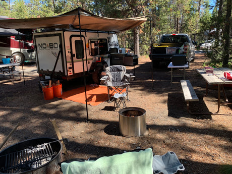 Relaxing RV rental campsite with a NO BO travel trailer, truck, fire pits, and outdoor seating under an awning.